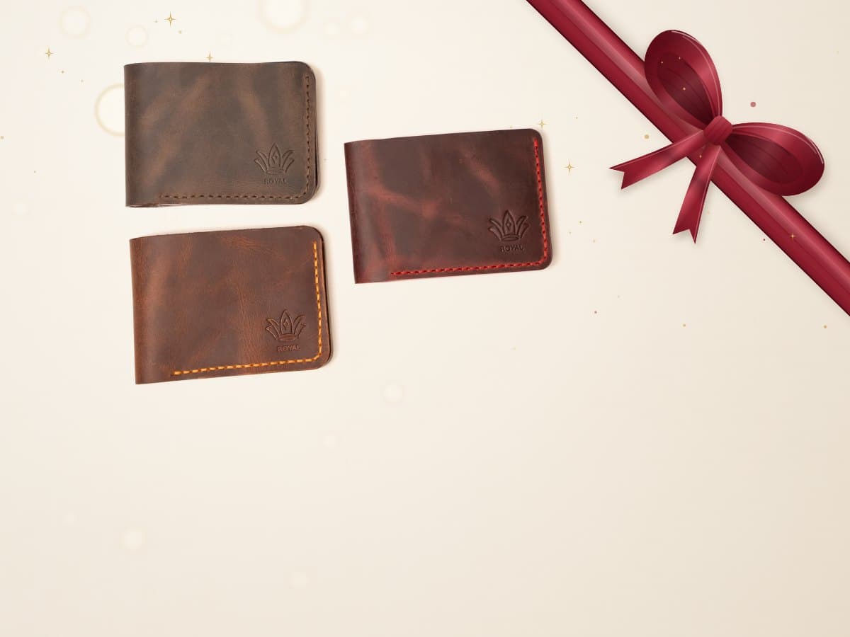 Three ROYAL leather bifold wallets in olive, tan and burgundy beside a cognac bifold standing open, on a warm cream surface